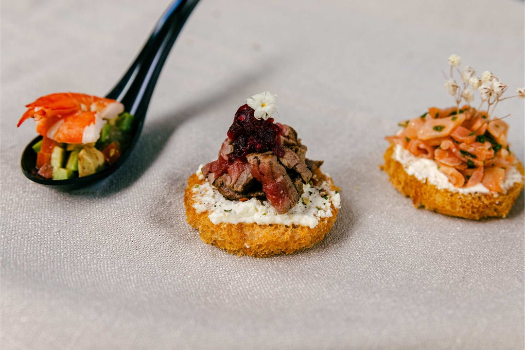 Three appetizer bites on a textured surface with a black spoon holding shrimp.
