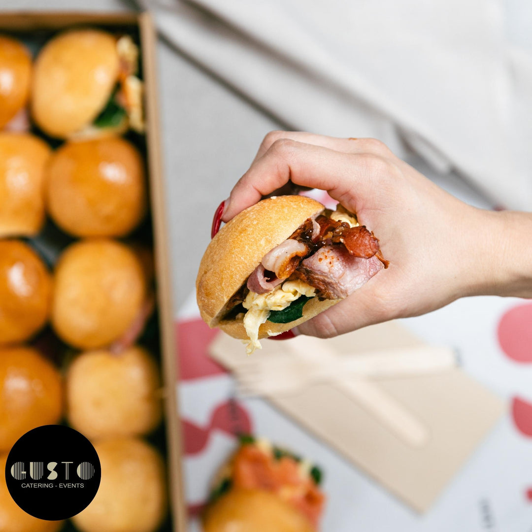 Hand holding a burger with a blurred background of sliders and a box.