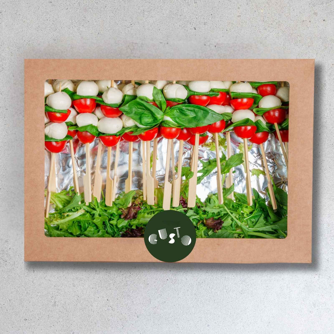 Packaged salad skewers with tomatoes, mozzarella, and basil in a brown box on a gray background.