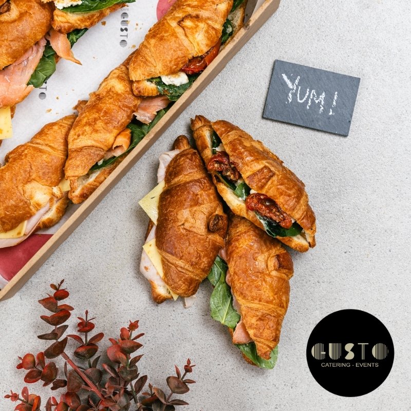 Croissant sandwiches on a gray surface with a 'YUM!' sign and Gusto Catering & Events logo.