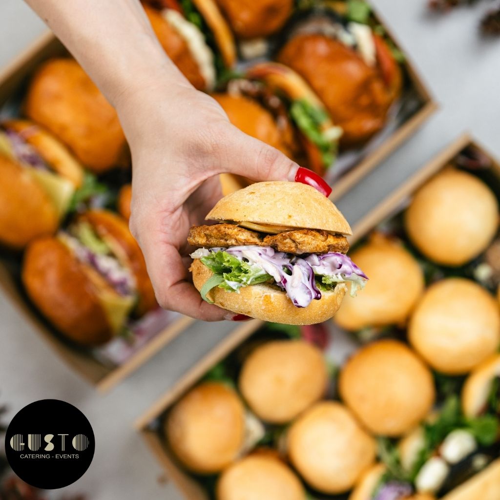 Hand holding a burger with visible ingredients, surrounded by more burgers in boxes, with 'Gusto Catering Events' logo.