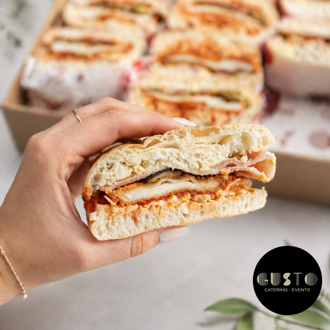 Hand holding a sandwich with visible layers of ingredients, with a blurred background of more sandwiches and 'Gusto Catering & Events' logo.