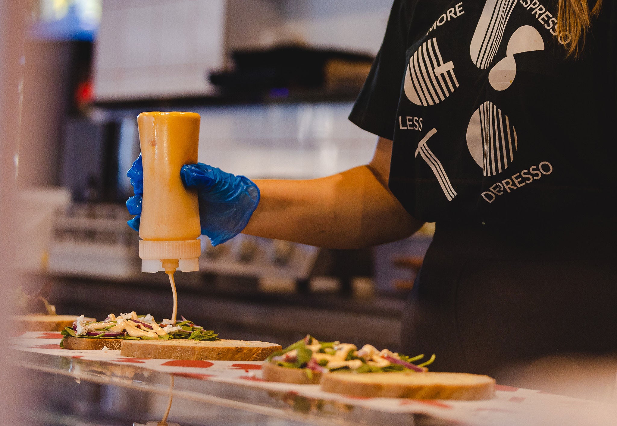 Person in a kitchen wearing gloves and a black shirt with 'Gusto' branding, pouring sauce onto food.