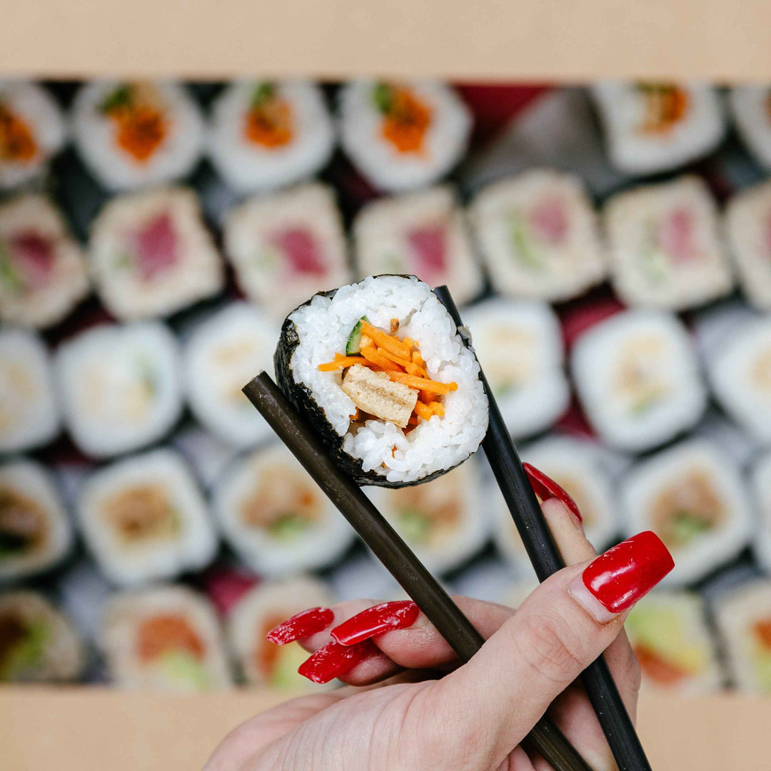Person holding a piece of sushi with chopsticks in front of a display of sushi rolls.